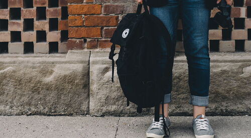 Person steht vor einer Backsteinwand auf einem Gehweg, trägt Jeans und graue Turnschuhe und hält einen schwarzen Rucksack in der rechten Hand.