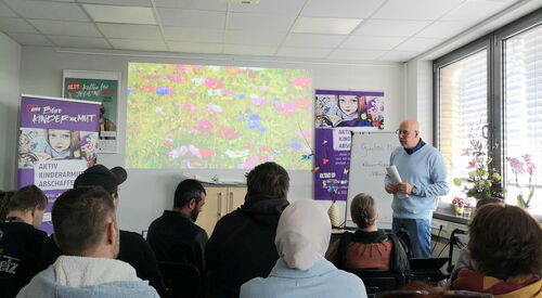 Großer Raum. Teilnehmende sitzen in Stuhlreihen, vorne steht ein Mann rechts am Fenster.