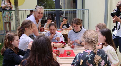 Draußen vor einem Gebäude. Kinder sitzen an Tischen und essen gut gelaunt Erdbeeren. Eine blonde Frau mit blonden Haaren und Brille auf den Kopf geschoben, steht am vorderen Tisch und lächelt.