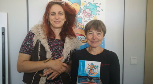 2 Frauen beide lächeln in die Kamera. Rechts kleine ältere Frau mit kurzen Haareb hält eine Spendendose mit Wellenreiter Logo in der Hand.