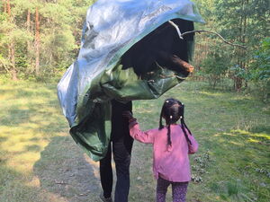 Zwei Personen gehen auf einem Waldweg. Eine größere Person trägt eine zusammengerollte, bunte Plane über der Schulter, neben ihr läuft ein Kind mit pinker Jacke.