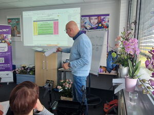 Mann erklärt Präsentation die an der Wand läuft. Auf dem Fensterbrett rechts stehen Orchideen.