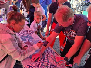 Auf einem Unterarm wird mit einer Schablone und Farbe ein Tattoo-Motiv aufgesprüht. Einige Leute auch Kinder stehen am Stand und sehen zu.