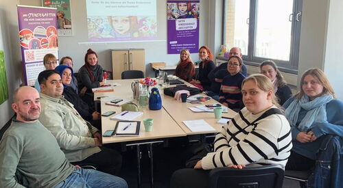 Großer Konferenzraum des Büro Kindermut. 13 Personen sitzen um einen großen Tisch und sehen in die Kamera. An der hinteren Wand läut eine Präsentation.