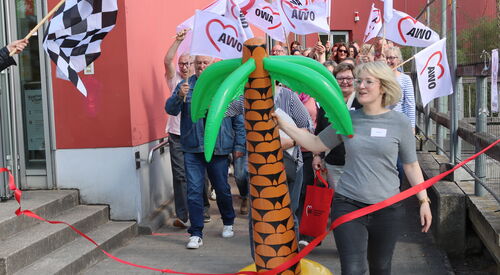 Eine Zielflagge links im Bild. Eine blonde Frau mit einer großen Gummi-Palme tritt über die Ziellinie (rotes Band) Hinter hier Teilnehmende im weißen AWO Flaggen.