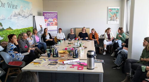 Großer heller Raum. Viele Menschen sitzen in um großen Tisch. Auf dem Tisch Getränke.