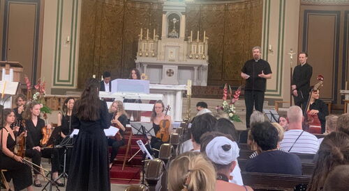 In einer Kirche. Junge Menschen mit ihren Intrumenten in festlicher schwarzer kleidung. Dirigentin von hinten mit langen dunklen Haaren.