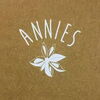 Weißer Schriftzug „ANNIES“ in Rundbogenform über einer stilisierten weißen Blüte auf braunem Hintergrund.