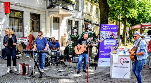 Band mit Musikinstrumenten vor einem AWO Stand. Eine Frau am Gesang, 4 Männer an Gitarre, Bass, Schlagzeug und Keyboard an einer Straßenecke in einer Fußgängerzone.