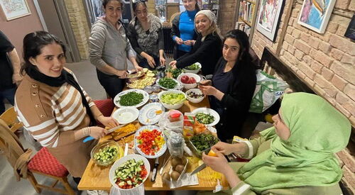 Raum mit Steinmauern und Bildern an der Wand. Voller Tisch, Frauen bei der Zubereitung einer Mahlzeit. Gutgelaunte Gemeinschaft.