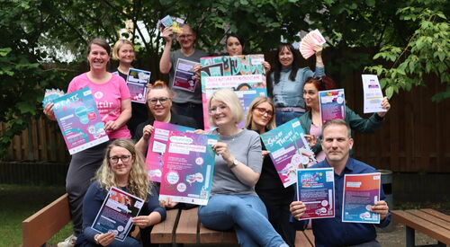 Gruppenbild: Menschen von AWO, Kultür und anderen Einrichtungen, Sitzen beziehungsweise stehen auf Tisch und bänken. Sie halten Plakate und Flyer und sind sehr fröhlich.