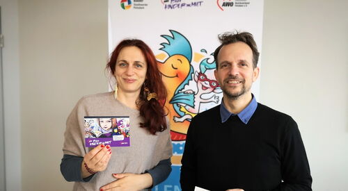 Eine Frau mit roten langen Haaren und ein Mann stehen nebeneinander und lächeln in die Kamera. Die Frau hält eine Karte vom Büro kindermut, der Mann eine karte vom Wellenreiter.