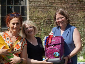 3 Frauen lachen fröhlich in die Kamera. Eine mit einer gelb grünen Schultüte in dem Armen, die beiden anderen halten einen lila Schulranzen.