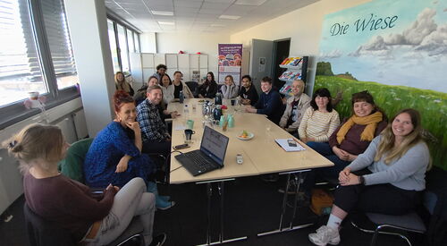 Großer Beratungsraum 16 Menschen sitzen um großen Tisch. Rechts an großes Bild einer Wiese mit blauen Himmel an der Wand.