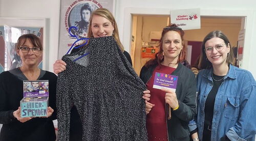 4 Frauen in einem Raum. Die 2. von links hält einen Kleiderbügel mit einem kurzen Kleid vor sich. Alle lächeln in die Kamera.