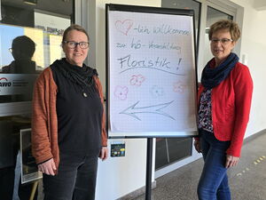 Zwei Frauen stehen im Flur, zwischen ihnen ein Flipchart mit Wegweiser-Pfeil zur Info-Veranstaltung Floristik.