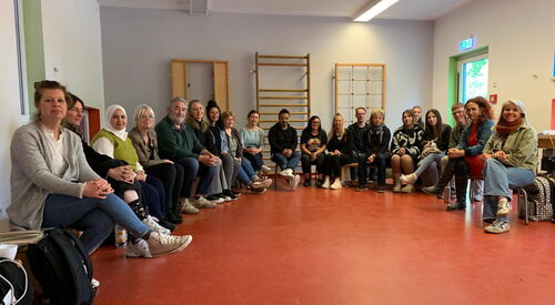 Gruppenfoto der Teilnehmenden. Sitzen in großem Halbkreis in einer Turnhalle/Sportraum.