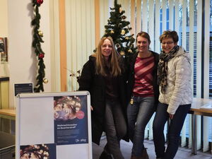 Weihnachtlich geschmückter Raum. 3 Frauen lehnen sich an einen Tisch. Sie lächeln in die Kamera.
