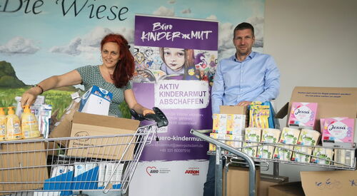 Im Büro Kinderarmut, eine Frau und ein Mann, jeweils mit einem vollen Einkaufswagen mit Hygieneartikeln zwischen ihnen das lila Roll-Up vom Büro Kinderarmut.