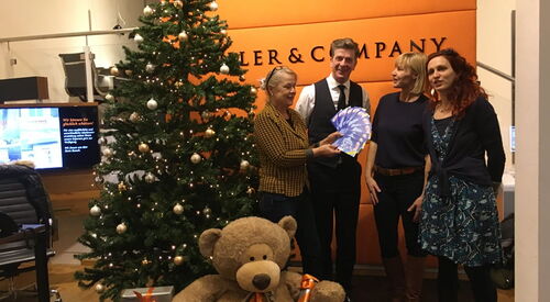 Ein Weihnachtsbaum, mit Geschenken und großem Teddy. Daneben stehen 4 Personen. Eine blonde Frau hält gefächert lila Karten/Gutscheine in der Hand.