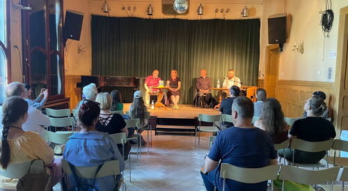 Auf einer Bühne sitzen 4 Personen im Rampenlicht, Davor das Publikum. Von hinten Richtung Bühne fotografiert.