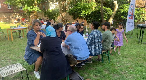Draußen, Sonne scheint. Tische stehen im Freien im Schatten, Menschen, Kinder mit unterschiedlichen Hautfarben sitzen auf Bänken und essen und trinken.