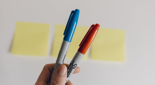 Eine Hand hält 2 Stifte hoch, dahinter 3 Post-its auf einen weißen Tisch geklebt.