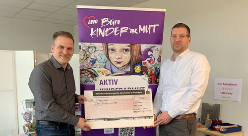 2 Männer halten in der Mitte einen Spendencheck. Das Lila Büro Kindermut Roll-Up steht zwischen ihnen.