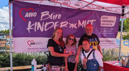 Gruppenbild: 3 Frauen und ein Mann. In einem Pavillon mit Banner vom Büro Kindermut. Sie lächeln fröhlich in die Kamera.