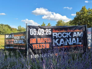 Schwarzes Veranstaltungsbanner mit der Aufschrift „Rock am Kanal“ hängt an einem Zaun, im Hintergrund grüne Bäume und blauer Himmel.