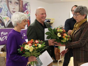 Eine ältere Frau mit lila Pullover mit großem Blumenstrauß und einem Dankesschreiben in den Händen. Neben ihr ein älterer Mann der einen Blumenstrauß von einer Frau überreicht bekommt. Ein feierlicher Anlass.