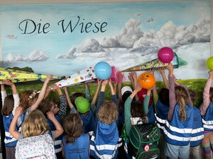 Kinder stehen mit dem Gesicht zur Wand und strecken Zuckertüten und Ballons nach oben. Sie stehen vor einem Bild mit Titel: Die Wiese