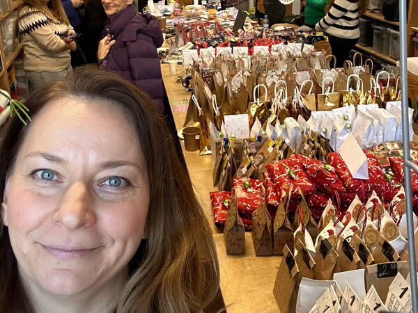 Selfie Optik. Frau mit braunen langen Haaren. Auf dem Tisch rchts neben ihr viele kleine unterschiedliche Geschenktüten und Päckchen. Personen in Hintergrund.