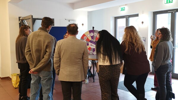 Großer Raum, mit Fließen Boden. Eine kleine Gruppe bildet einen Halbkreis um eine Art Glücksrad.