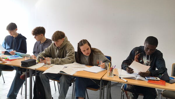 5 Jugendliche mit Schulmaterialien sitzen nebeneinander an aneinander gereihten Tischen.