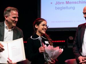 Frau mit Blumen und Mikrofon in der Hand. Sie lächelt und freut sich über die Auszeichnung. Links und rechts von ihr zwei Männer. In weißem Hamd und dunklem Sakko.