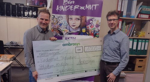 Zwei Männer mit einem großen Spendenscheck zwischen sich. Beide lächeln in die Kamera. Sie stehen in einem Büroraum.