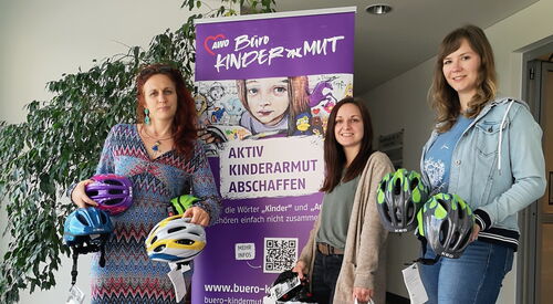 Drei Frauen mit Fahrradhelmen und Tüten mit dem Logo vom Radhaus stehen in einem Flur. Hinter ihnen ein lila Aufsteller vom Büro Kindermut.