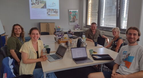 3 Frauen, 2 Männer an einem Tisch in einem Konferenzraum. An der Wand eine Präsentation. Die 5 Menschen schauen in die Kamera und lächeln.