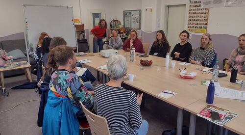 Besprechung: großer Raum. 12 Menschen sitzen um einen Tisch. Eine Frau it rotem Pulli steht.