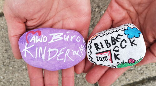 Zwei Hände auf denen jeweils ein bemalter Stein liegt. links: lila Stein mit weißer Schrift: AWO Büro Kindermut   rechts: weißer Stein mit schwarzer Schrift: Ribbeck 2025 mit Wolke und Wiese.