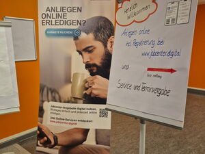 Plakat und Flipchart. Flipchart mit Text: Herzlich willkommen. Anliegen online incl. Registrierung bei jobcenter digital     und roter Pfeil nach rechts