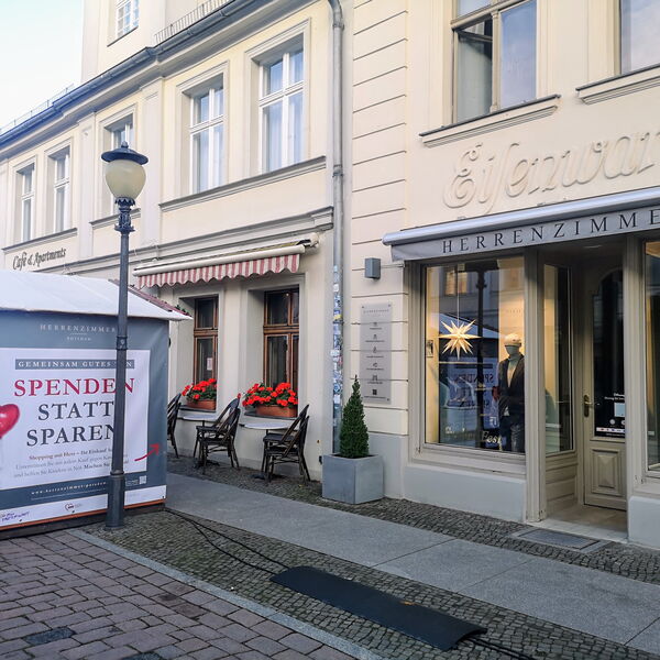 Ladengeschäft in Einkaufsstraße. Herrenzimmer. Links davon eine Laterne und eine kleine Bude mit dem Banner: Spenden statt sparen an der Außenwand.