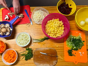 Tisch von oben. Darauf Schneidebretter, Schüsselchen mit geschnittenen Möhren, Tomaten, Zwiebeln, Champions und Kräutern.