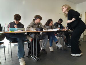 4 Jugendliche sitzen an aneinandergereihten Tischen nebeneinander. Auf den Tischen Schulmaterial. Ihnen gegenüber auf der innenseite des Tisches eine blonde Jugendliche stehend.