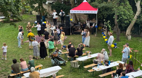 Von schräg-oben: Eine Wiese, mit Bänken und Tischen, eine kleine Bühne und roter Pavillon. Eine Band spielt auf der Bühne, Gäste tanzen davor.
