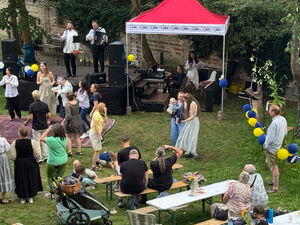 Von schräg-oben: Eine Wiese, mit Bänken und Tischen, eine kleine Bühne und roter Pavillon. Eine Band spielt auf der Bühne, Gäste tanzen davor.