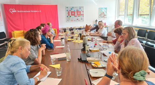 Großer Raum, große Tische. Viele Menschen sitzen sich gegenüber, einige machen sich Notizen.