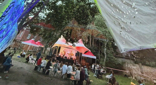 Festwiese mit Menschen und Pavillons. In den Bäumen sind farbige Tücher gespannt.