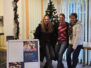 Weihnachtlich geschmückter Raum. 3 Frauen lehnen sich an einen Tisch. Sie lächeln in die Kamera.